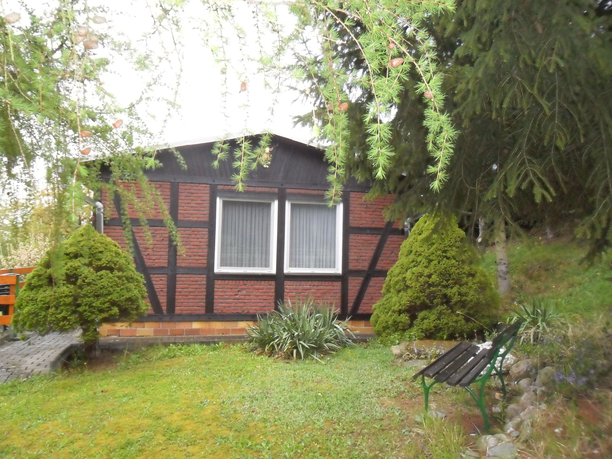 Der Bungalow von außen: Fachwerkfassade, gepflegter Rasen und eine Bank unter alten Bäumen