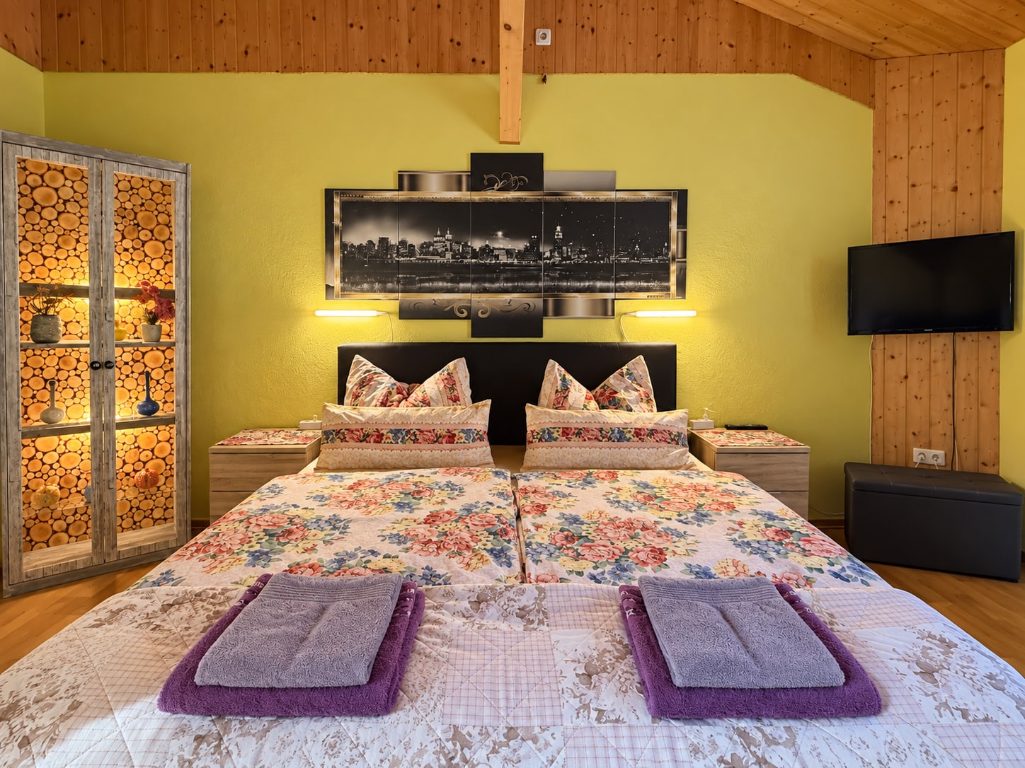 Doppelbett der Ferienwohnung mit floraler Bettwäsche und warm beleuchteter Vitrine
