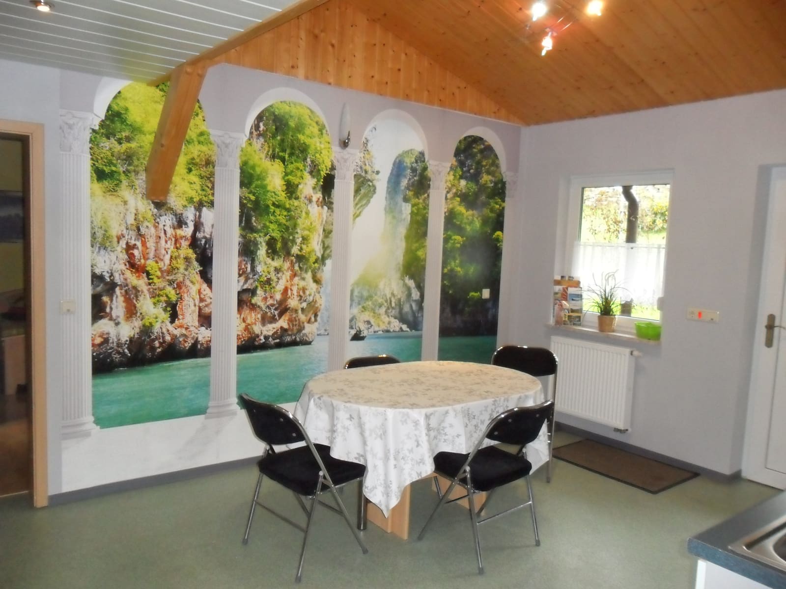 Essplatz der Ferienwohnung vor einer Fototapete mit Wasserfallmotiv, darüber eine helle Holzdecke