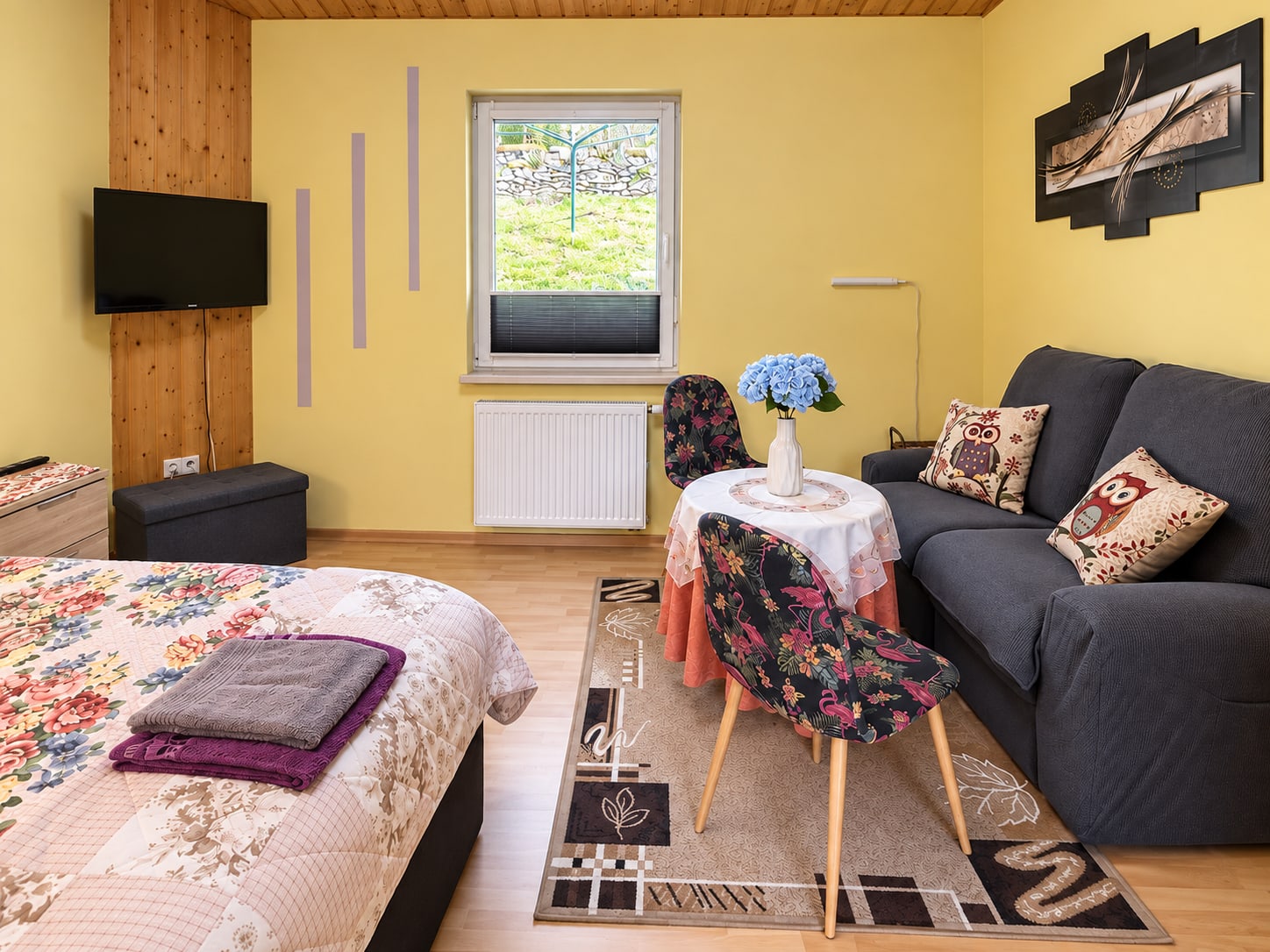 Heller Wohn- und Schlafraum der Ferienwohnung mit grauem Sofa, Doppelbett und gelben Wänden
